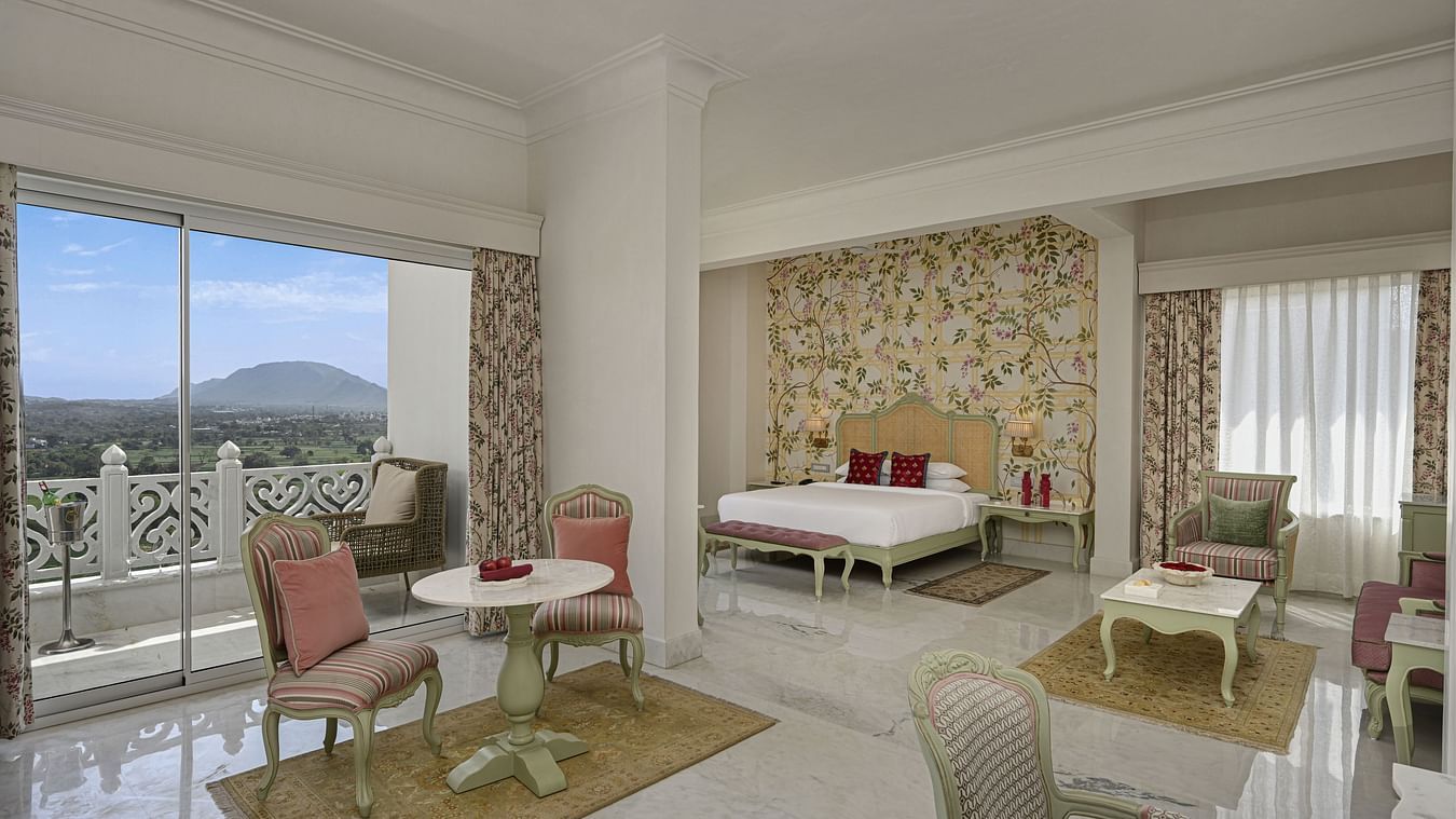 A bright and spacious Heritage Chamber Suite at Khas Mahal by Fateh Collection, Udaipur features a sitting area with pink chairs and a small table, a bed in the background, and a large window offering a view.
