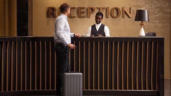 A guest with luggage stands at the hotel reception desk while a staff assists him.