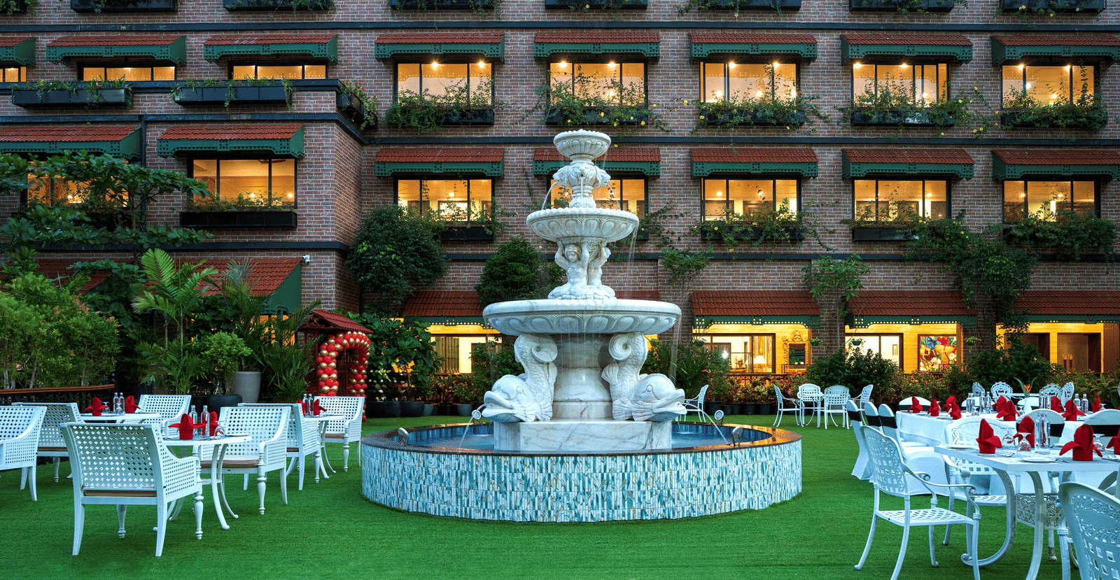 Outdoor dining area on a manicured lawn next to MAYFAIR Bay Resort, Paradeep's facade, set up with white tables and a fountain in the middle.