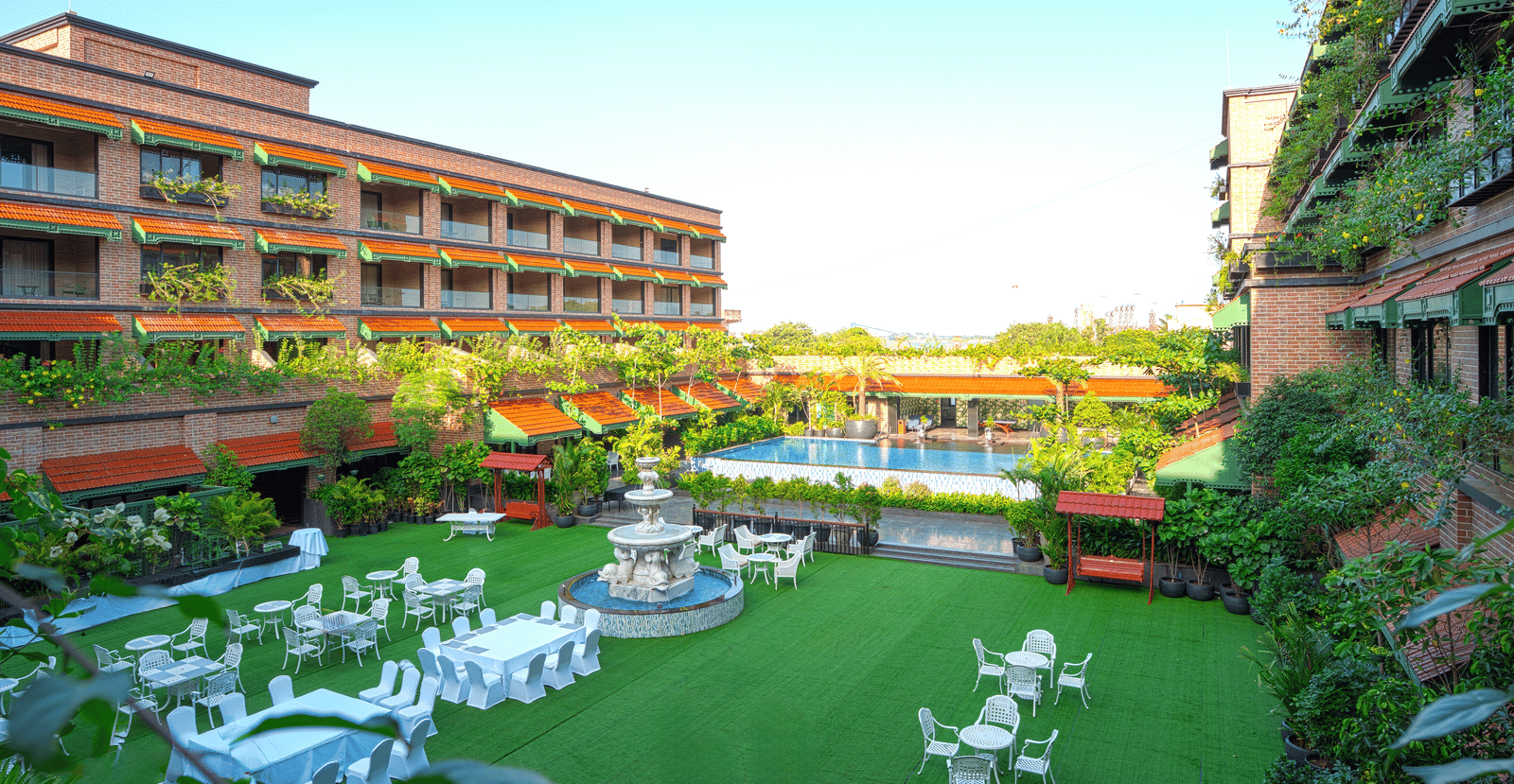 A wide view of the MAYFAIR Bay Resort, Paradeep, featuring a large lawn with white dining tables, a fountain, and a pool area nearby.