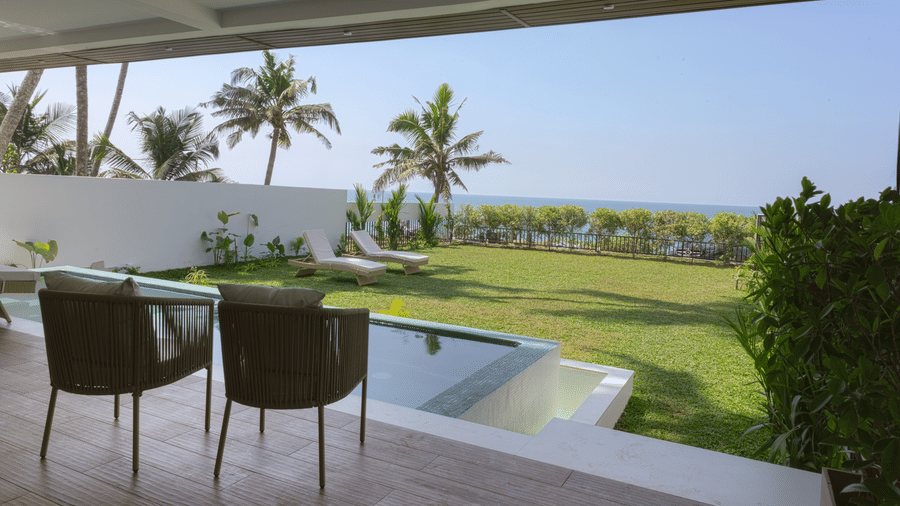 2 woven chairs sit on a covered patio at SUKAVI, overlooking a small pool, a green lawn, palm trees, and views of the ocean beyond.