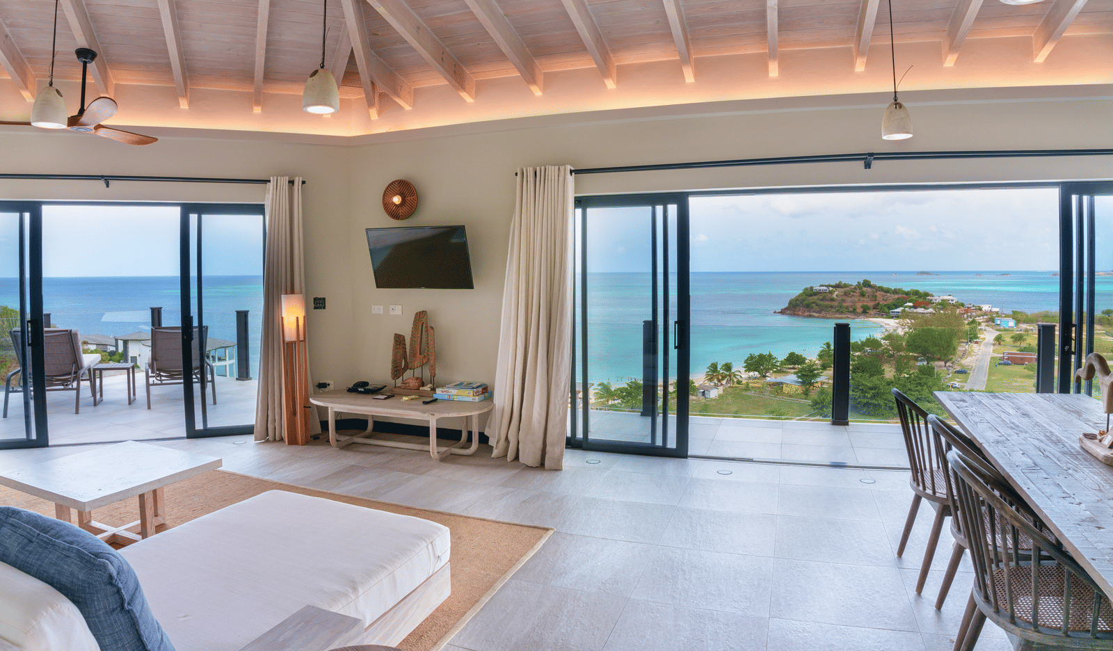 An open-plan living and dining area with a dining table, sofas, ceiling fans, and wide glass doors offering views of the swimming pool, sea, and coastline at Suite 35, Tamarind Hills Resort and Villas.