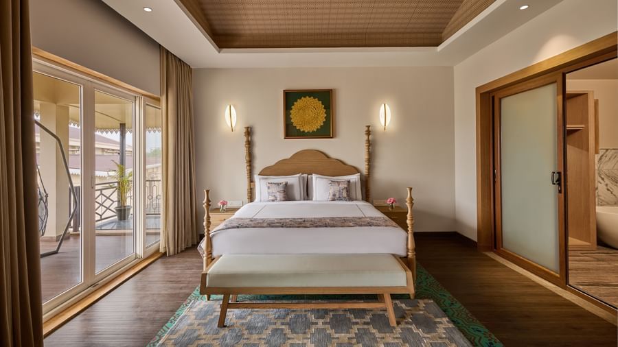 Elegant hotel bedroom with a wooden bed frame, white bedding, decorative rug, sliding glass doors leading to a balcony, and warm ambient lighting - Voyage The Solitaire Bliss.