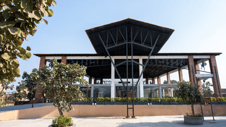 A front-facing exterior view featuring a modern building with a symmetrical dark roof structure, prominent brick pillars, and a low boundary wall with plants at Hotel Sonar Bangla, Ajodhya Hills.