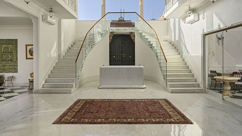 Interior view of a reception at Khas Mahal by Fateh Collection featuring a symmetrical, double staircase with white risers and stringers, glass railings, a dark central door, a brown rug on the floor, and natural light entering from a high window.