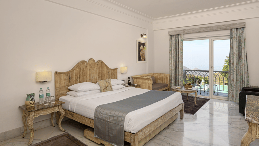 Superior Room with Balcony at Fateh Safari Resort, Kumbhalgarh featuring a double size bed, a bedside table with water bottles, a sofa with centre table, and a balcony with plunge pool with a view of the hill.