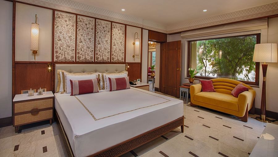 A beautifully designed bedroom with a plush king size bed, elegant floral touches, a cheerful yellow sofa, and a large window offering serene views of lush greenery - Heritage Village Resorts & Spa, Manesar.