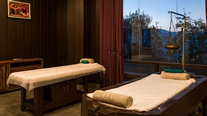 A tranquil spa room at Digantaa Resort in Mukteshwar featuring two massage beds covered in white sheets. The room features dark wood paneling and large windows with a view of greenery