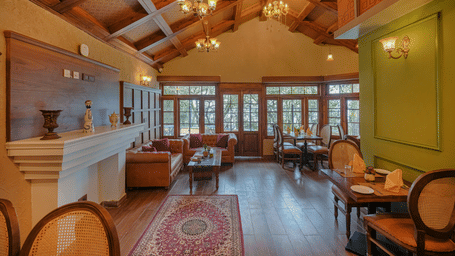 The well-lit Cafe Blusalzz, with wooden ceilings, sofas, chairs, decoratives placed on a shelf, chandeliers, large glass windows, and a door at BluSalzz Terrah Hills Resort, Dalhousie.