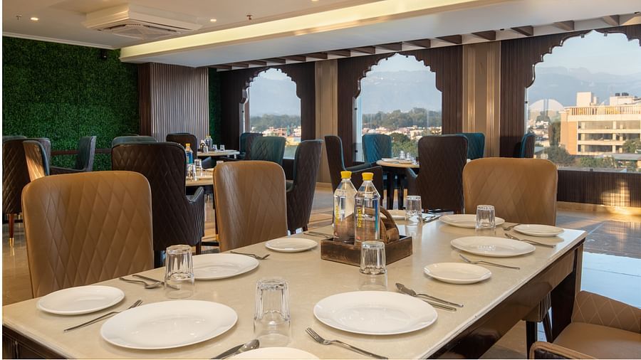 Image of multi-cuisine restaurant at DLS Grand Exotica in Dehradun featuring cutlery placed on the dining table and chairs are arranged with a scenic view of the city in the background