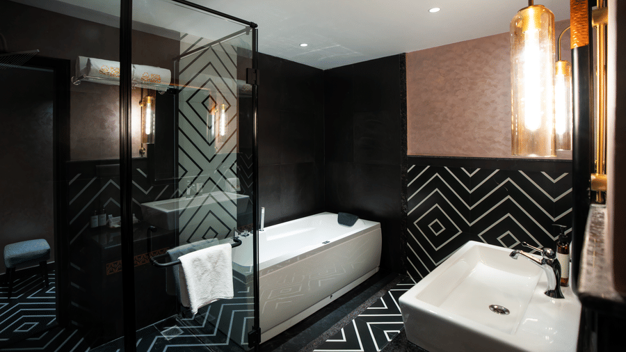 A luxurious bathroom in our Suite featuring a bathtub, glass shower enclosure, geometric wall patterns, and elegant pendant lighting beside the vanity area | Hotel Sonar Bangla, Susunia