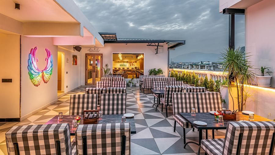 An outdoor rooftop dining area with checkered chairs, tables, and a view of the sky at dusk. It features a patterned floor, a painted mural on the wall, and a glimpse of the city - Lords Eco Inn Dehradun.
