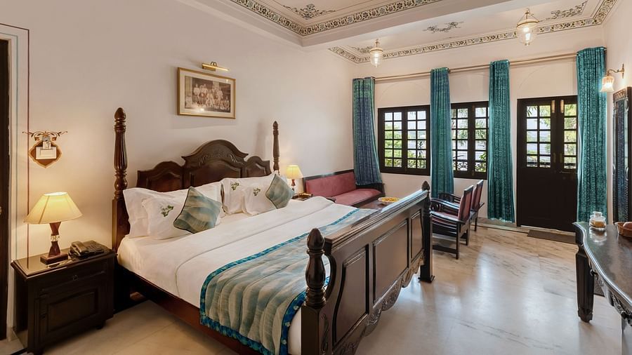 A spacious bedroom at Panna Vilas Palace, featuring a bedside table with a lamp and a series of windows to the side, with curtains pulled back | Panna Vilas Udaipur