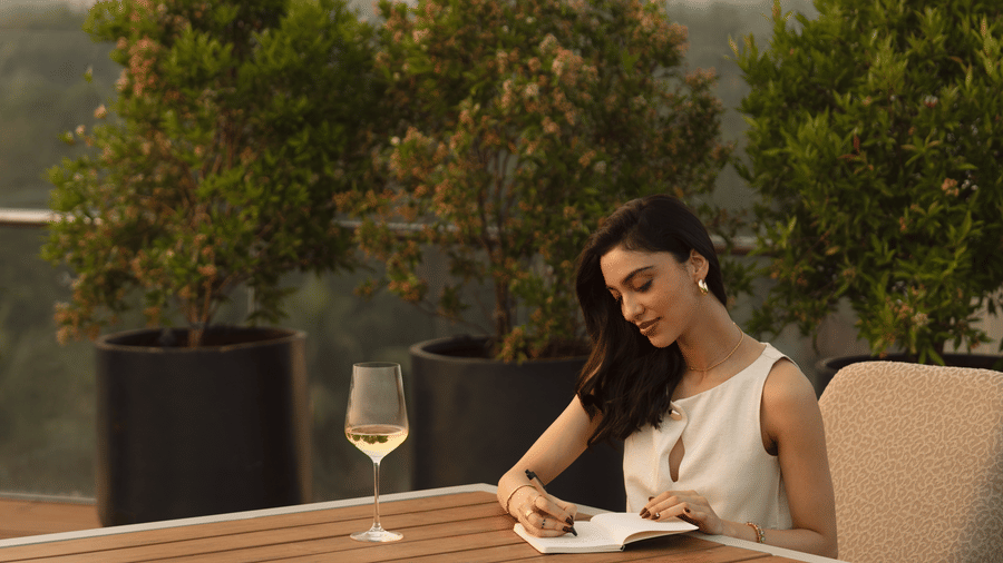 A woman seated at an outdoor table writing in a notebook, with a glass placed beside her and trees visible in the background at The Evren, Vagator.