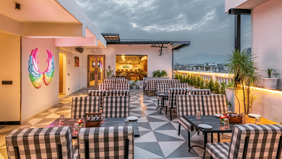 An elegant outdoor rooftop dining area with checkered chairs, tables, patterned floor, a vibrant mural on the wall and a view of the cityscape and clouded sky at the backdrop at Lords Eco Inn Dehradun.