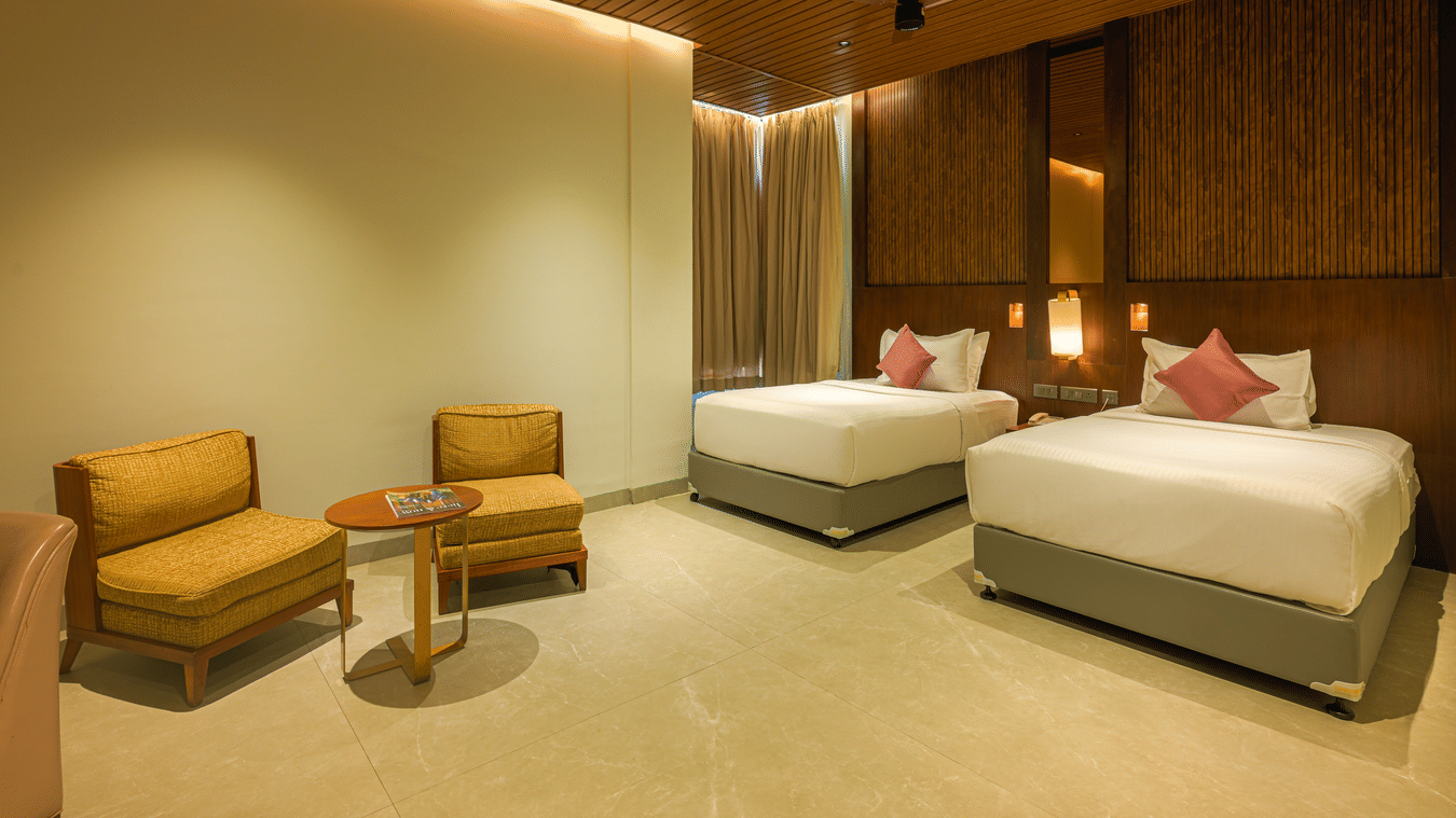 Hotel bedroom with two single beds, upholstered headboards, lounge chairs, wooden panelling and warm ceiling lighting.
