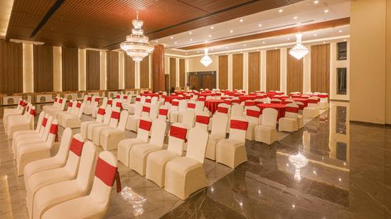 Spacious banquet hall featuring rows of white chairs with red accents, round tables, chandeliers and warm ambient lighting.