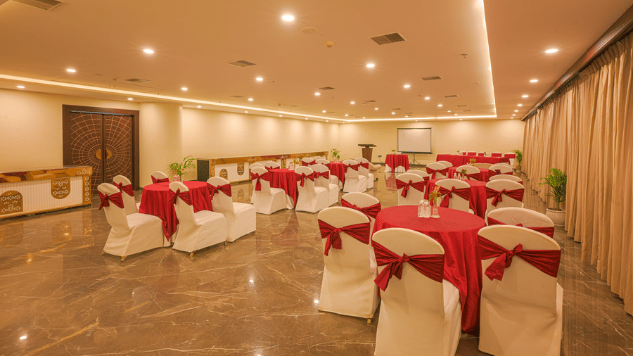 Banquet hall arranged with round tables, white and red covered chairs, floral centrepieces and warm ceiling lights.