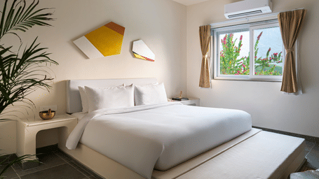 A white bedroom in a serene pool villa in Udaipur, featuring a double bed with white linen, an air-conditioning unit, a window, and wall artwork at Vihara.