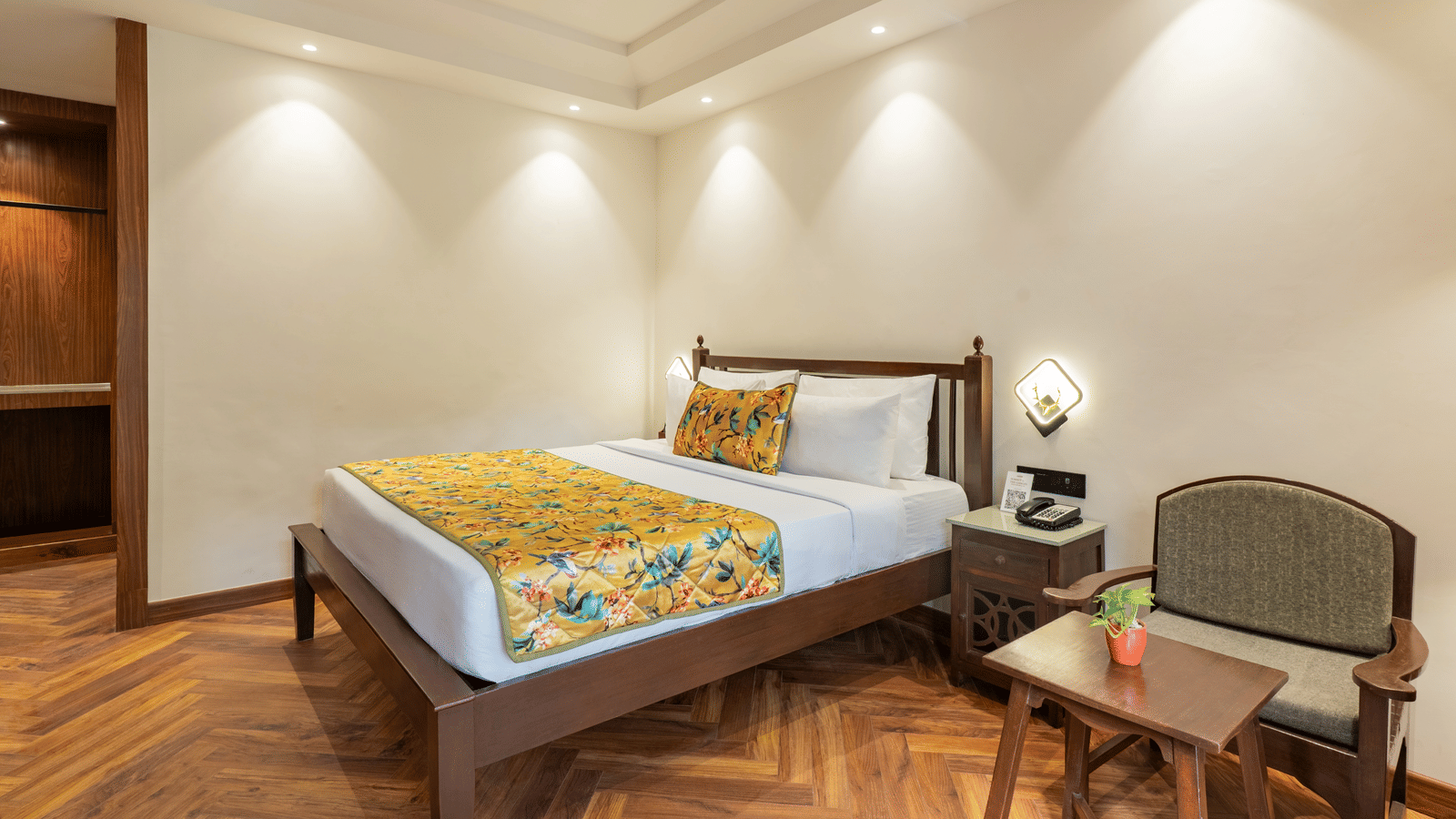 A well-lit bedroom featuring a wooden floor, a bed, a landline phone atop a small bedside cabinet, a chair facing a wooden table, and a small potted plant | Summit by the Ganges Beach Resort & Spa, Rishikesh