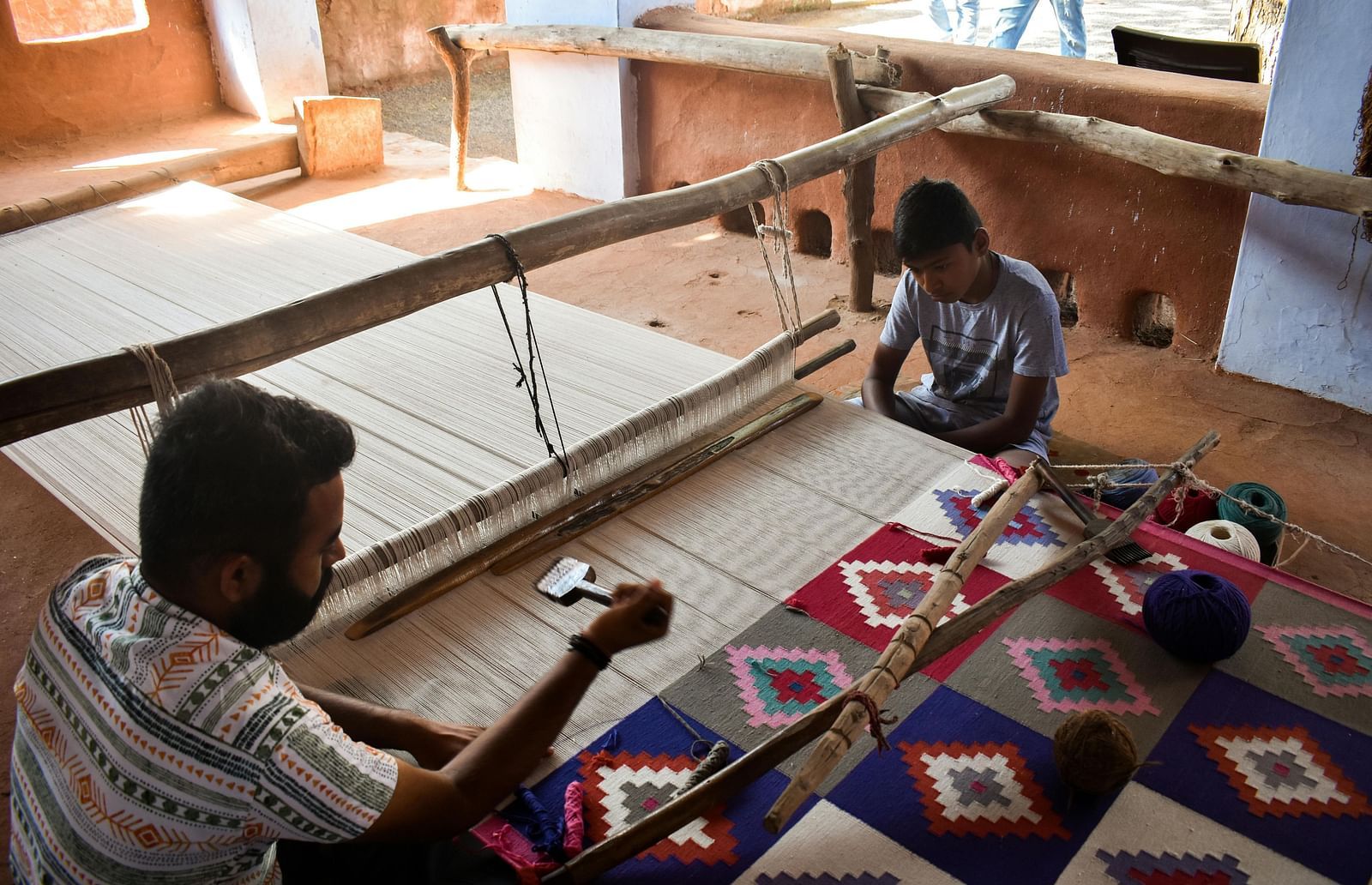 Two individuals seated on the floor, working on a traditional weaving loom