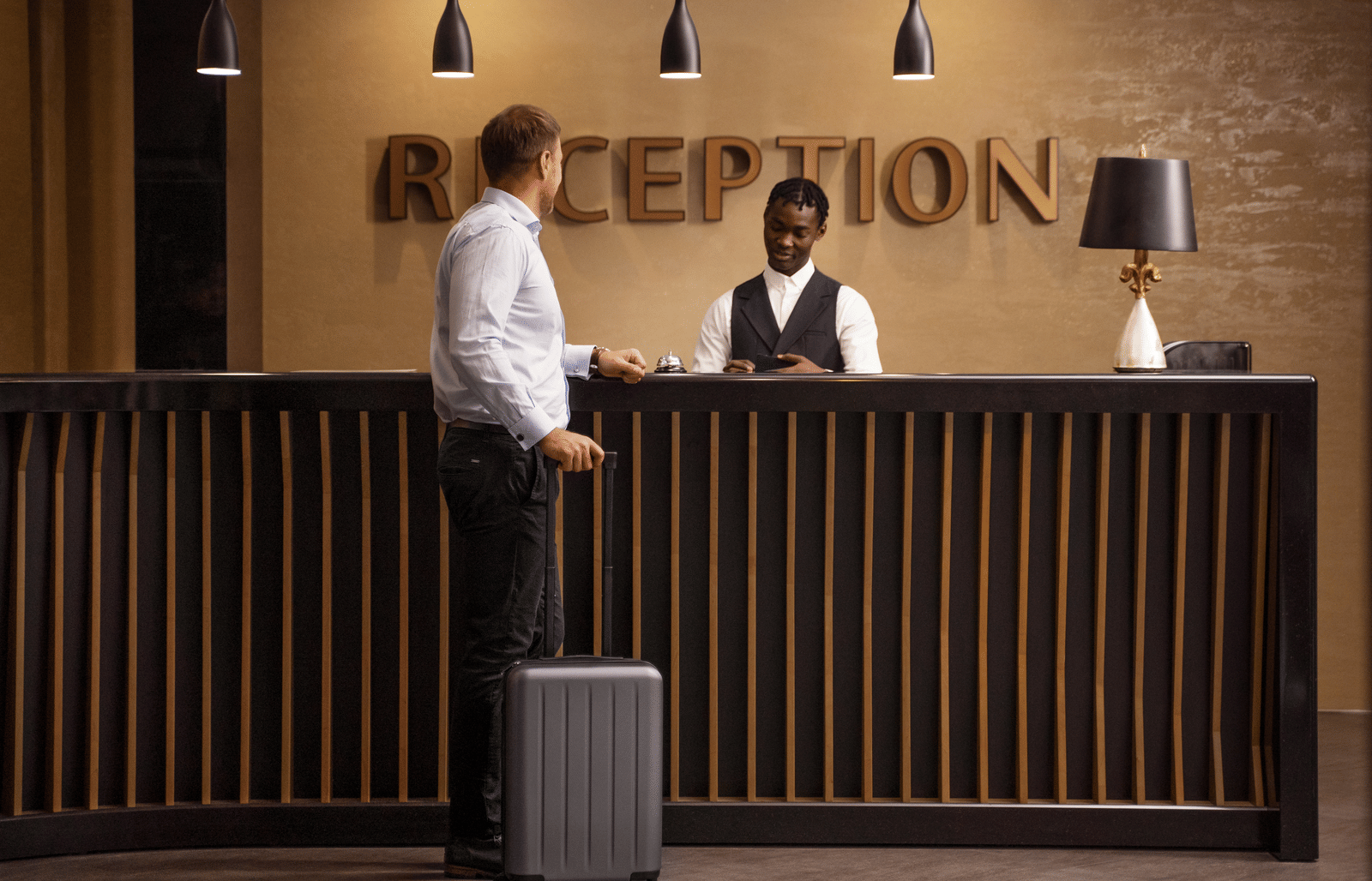 A guest with luggage stands at the hotel reception desk while a staff assists him.