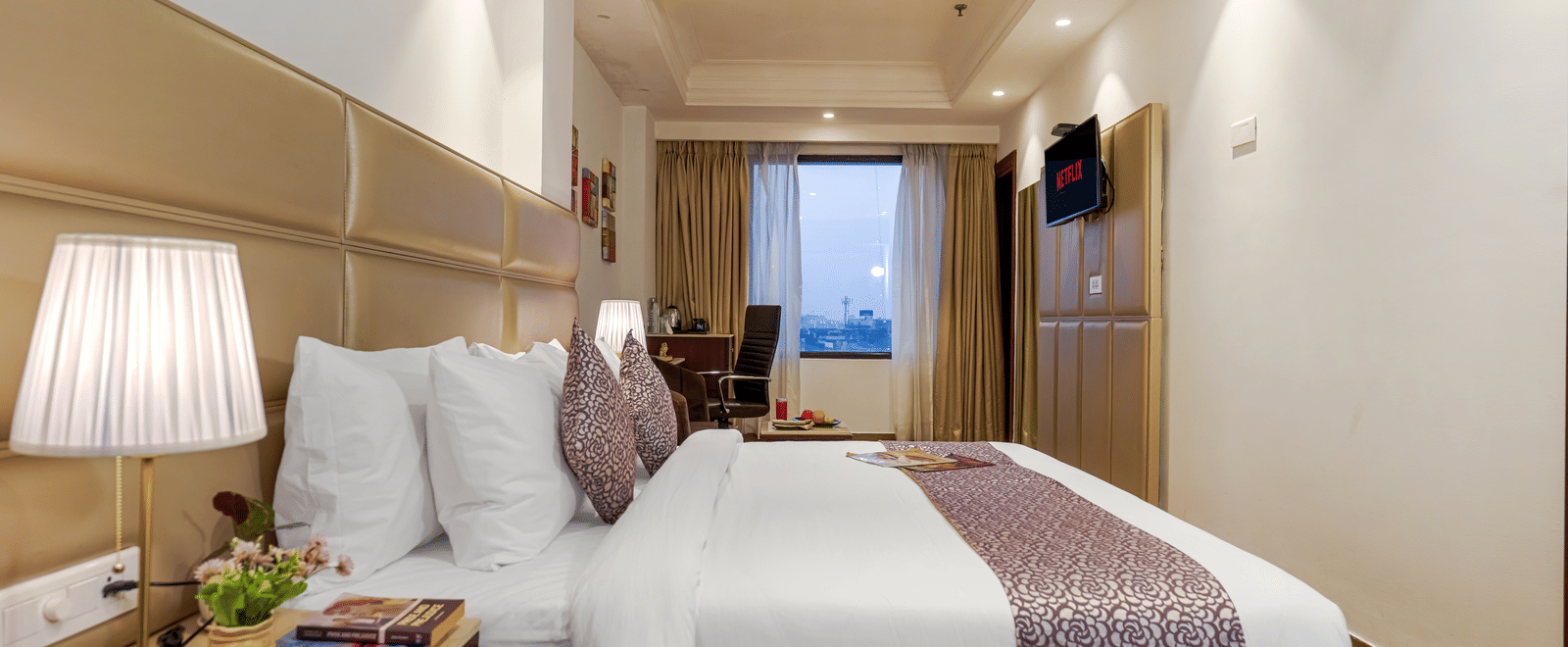 A modern, elegant hotel bedroom featuring a large bed with a patterned purple runner and a view of the city outside at BluSalzz Select City Centre, Amritsar.