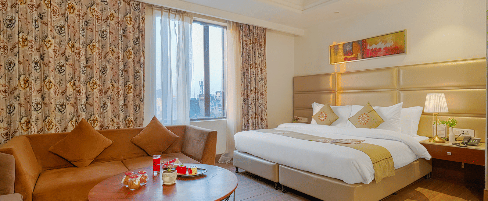 A long view of a hotel suite showcasing the bed, a TV area, desk, sofa, and window with patterned curtains at BluSalzz Select City Centre, Amritsar.