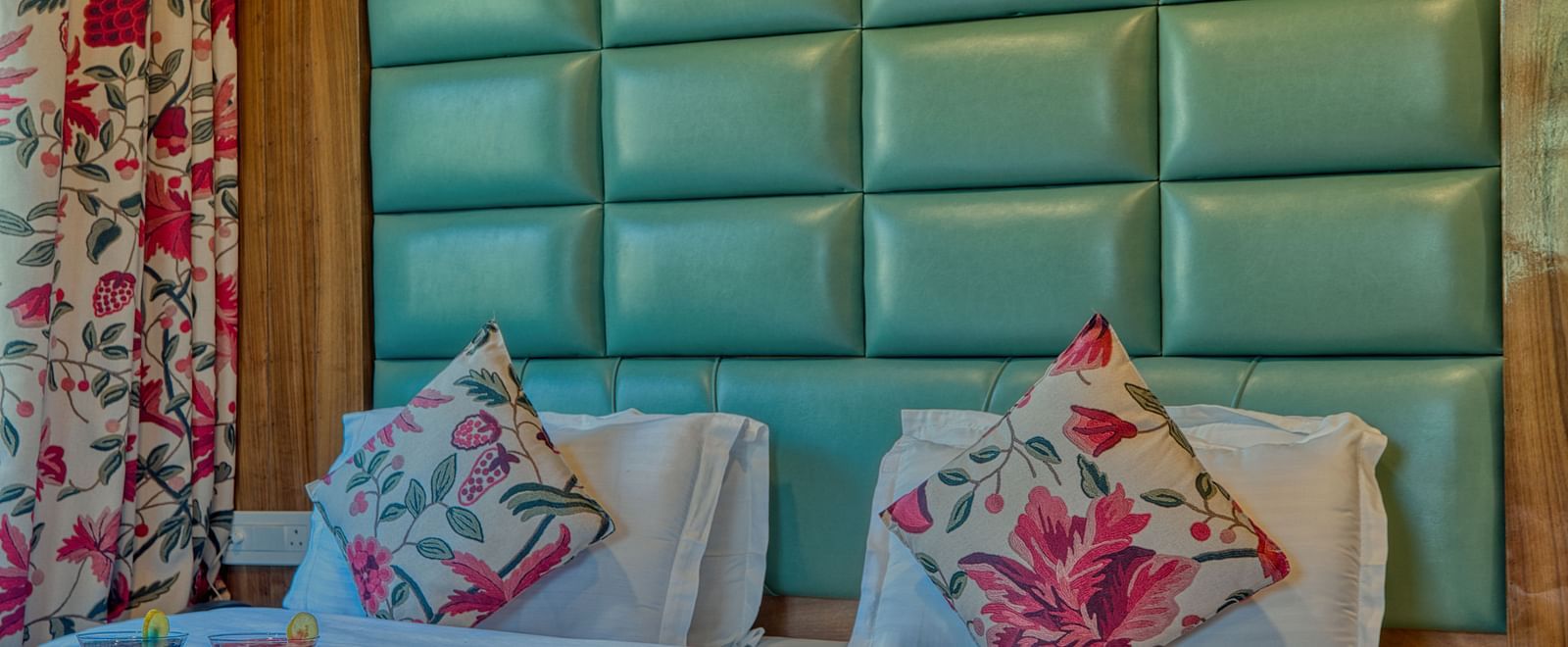 A close-up of a double bed at Royal Comfort Regency with a large padded green headboard, floral curtains and decorative cushions.