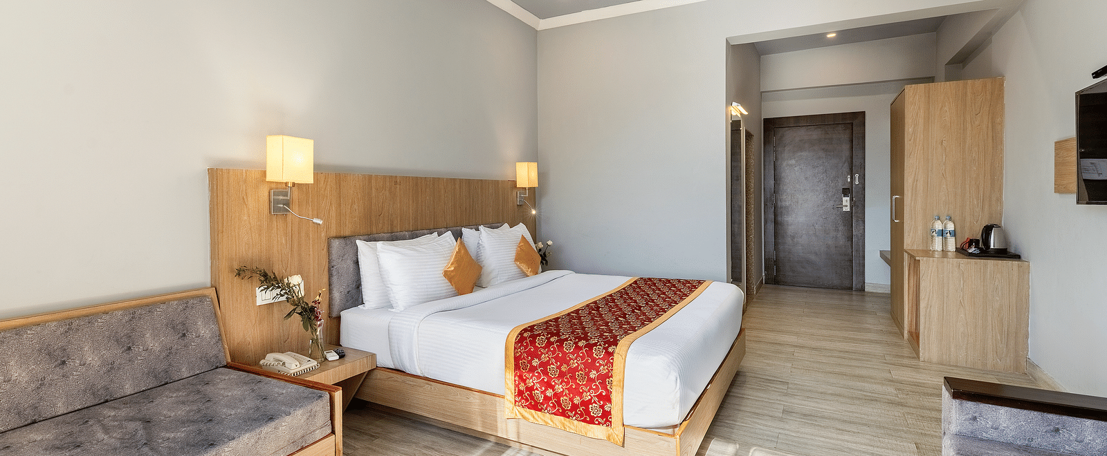 A tidy hotel room featuring a neatly made bed with a red runner, a gray sofa and a wooden side table with a fruit bowl on top of it - Via Lakhela Resort & Spa, Kumbhalgarh