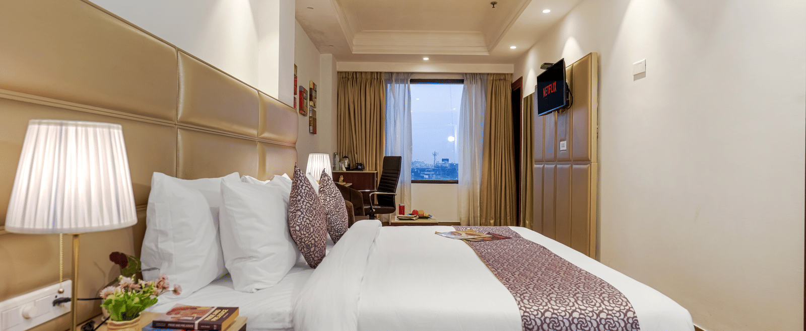 A modern, elegant hotel bedroom featuring a large bed with a patterned purple runner and a view of the city outside at BluSalzz Select City Centre, Amritsar.