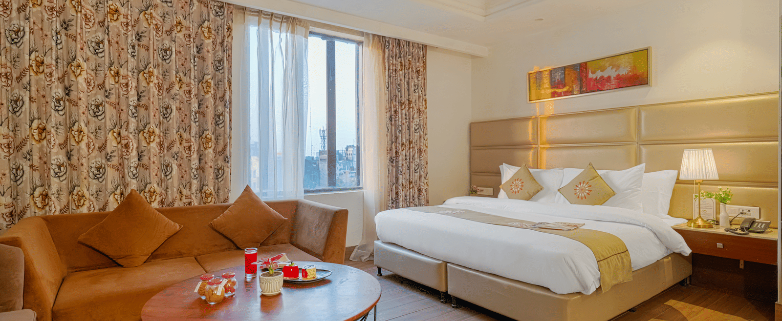 A long view of a hotel suite showcasing the bed, a TV area, desk, sofa, and window with patterned curtains at BluSalzz Select City Centre, Amritsar.