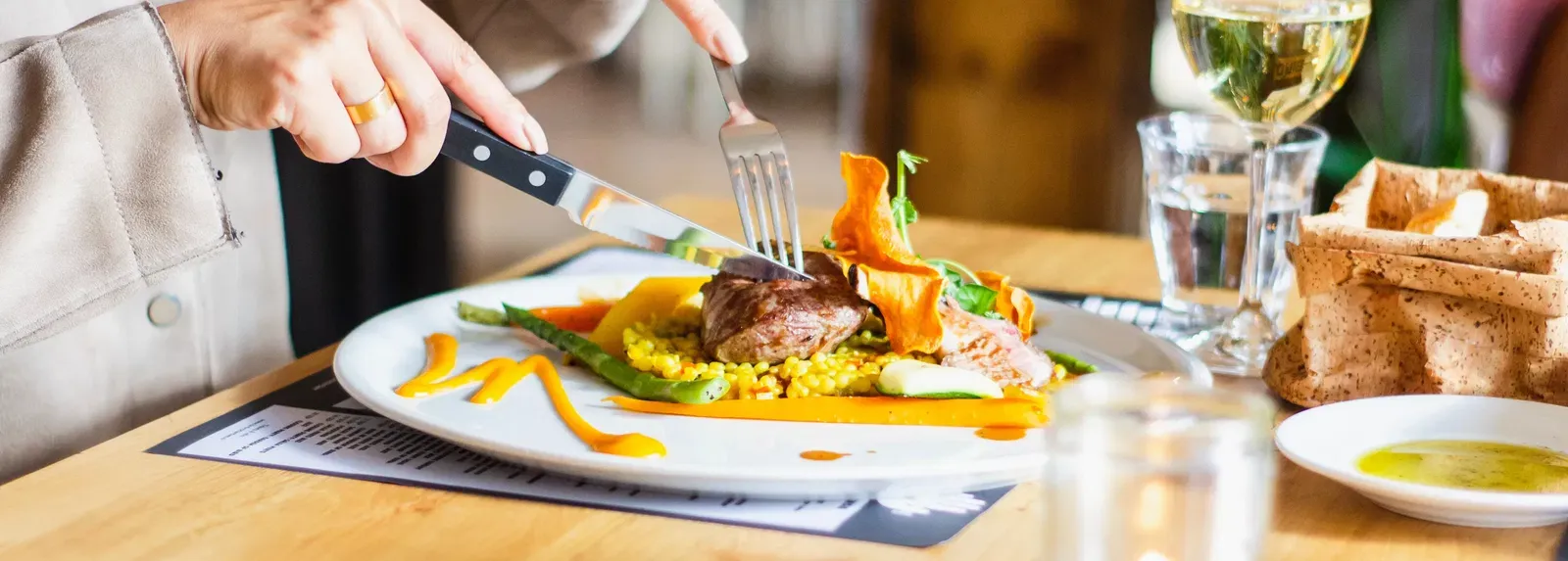 A person cuts into a gourmet meal featuring meat and vegetables on a white plate, accompanied by a glass of white wine and a lit candle on a wooden table.