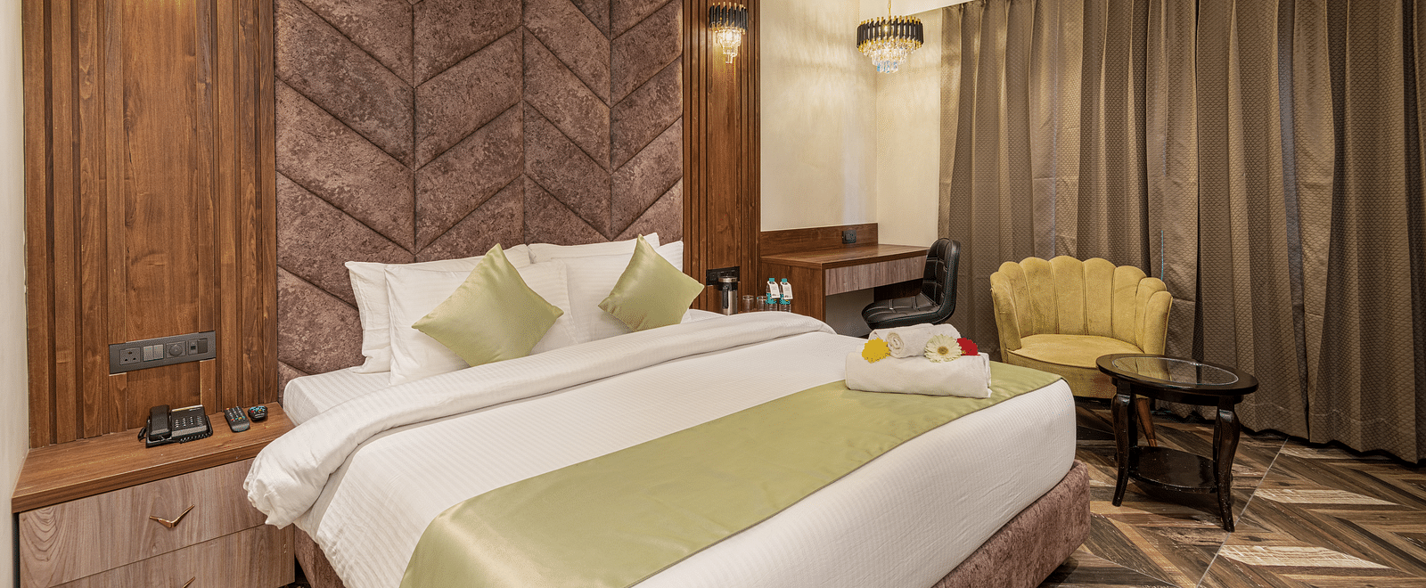Deluxe Room at DLS Grand Luxotica, Dehradun showcasing a plush bed with a nightstand next to it and a sitting space on the other side next to the window with a couch and coffee table