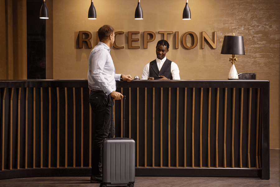 A guest with luggage stands at the hotel reception desk while a staff assists him.