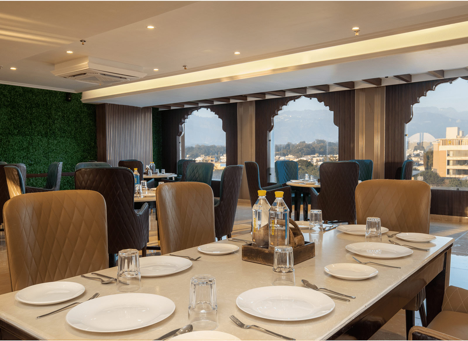 Image of multi-cuisine restaurant at DLS Grand Exotica in Dehradun featuring cutlery placed on the dining table and chairs are arranged with a scenic view of the city in the background