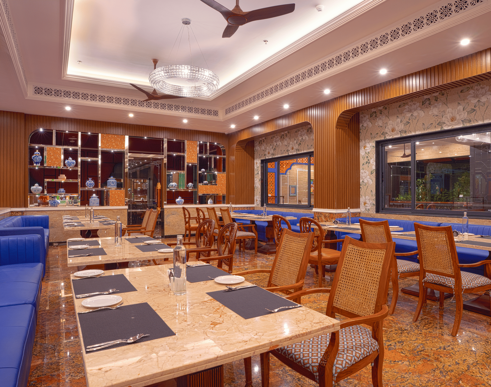 Brightly lit dining hall with wooden chairs, blue sofas, marble table, marble flooring, fans and  large window at MAYFAIR Bay Resort, Paradeep.