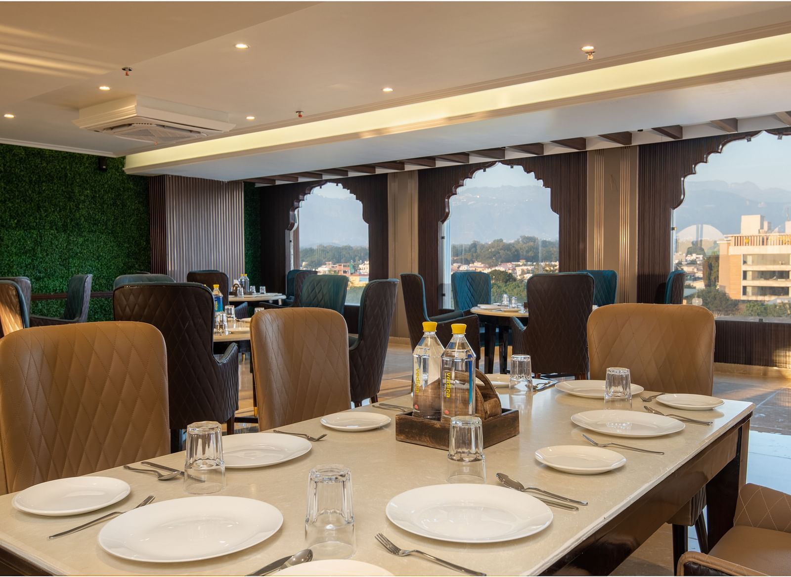 Image of multi-cuisine restaurant at DLS Grand Exotica in Dehradun featuring cutlery placed on the dining table and chairs are arranged with a scenic view of the city in the background