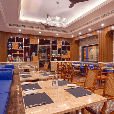 Brightly lit dining hall with wooden chairs, blue sofas, marble table, marble flooring, fans and  large window at MAYFAIR Bay Resort, Paradeep.