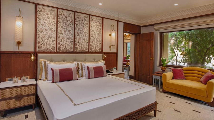 A beautifully designed bedroom with a plush king size bed, elegant floral touches, a cheerful yellow sofa, and a large window offering serene views of lush greenery - Heritage Village Resorts & Spa, Manesar.