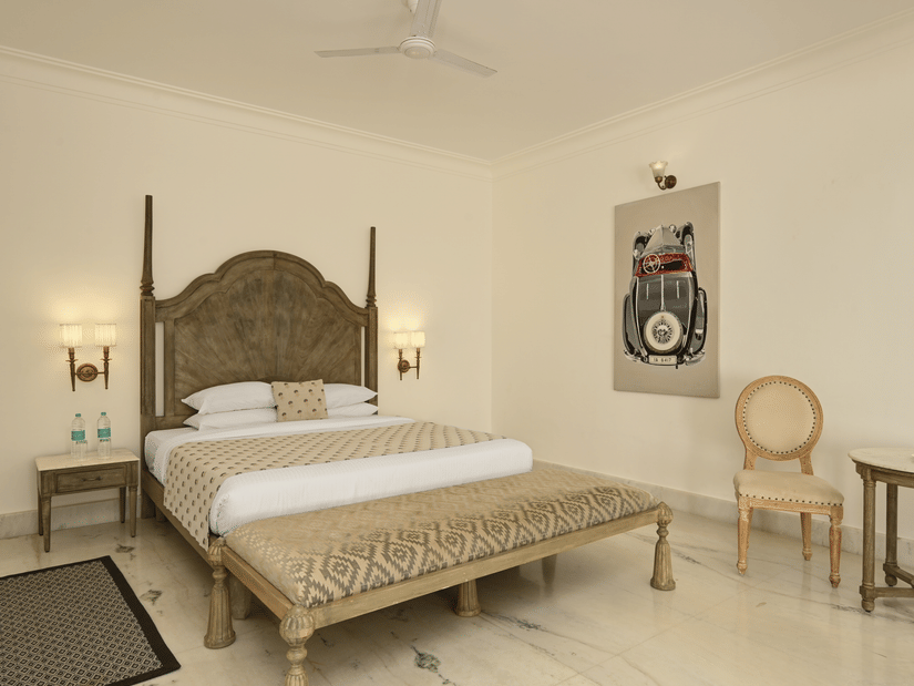 Bedroom of Superior Suites at Fateh Safari Suites, Kumbhalgarh featuring double size bed, a sofa, bedside table with lights on either side, a wooden chair, and a table.