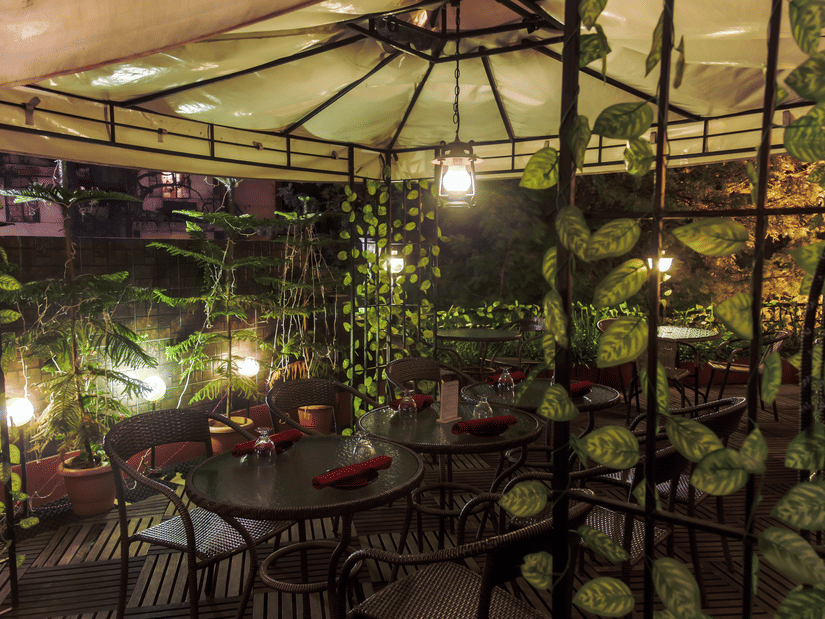 Outdoor restaurant at Polo Towers, Shillong with wicker chairs, round tables set with red napkins under a canopy hung with leafy vines and lanterns.