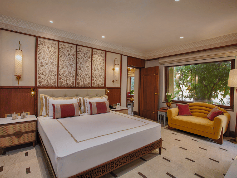 A bedroom with a king size bed, elegant floral touches, a cheerful yellow sofa, and a large window offering serene views of lush greenery | Heritage Village Resorts & Spa, Manesar.