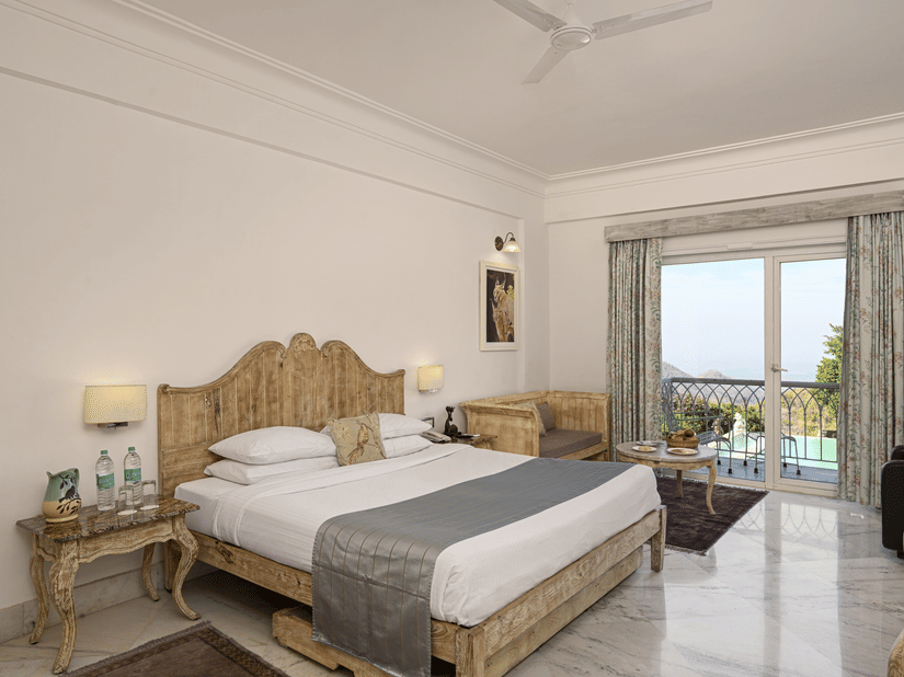 Superior Room with Balcony at Fateh Safari Resort, Kumbhalgarh featuring a double size bed, a bedside table with water bottles, a sofa with centre table, and a balcony with plunge pool with a view of the hill.