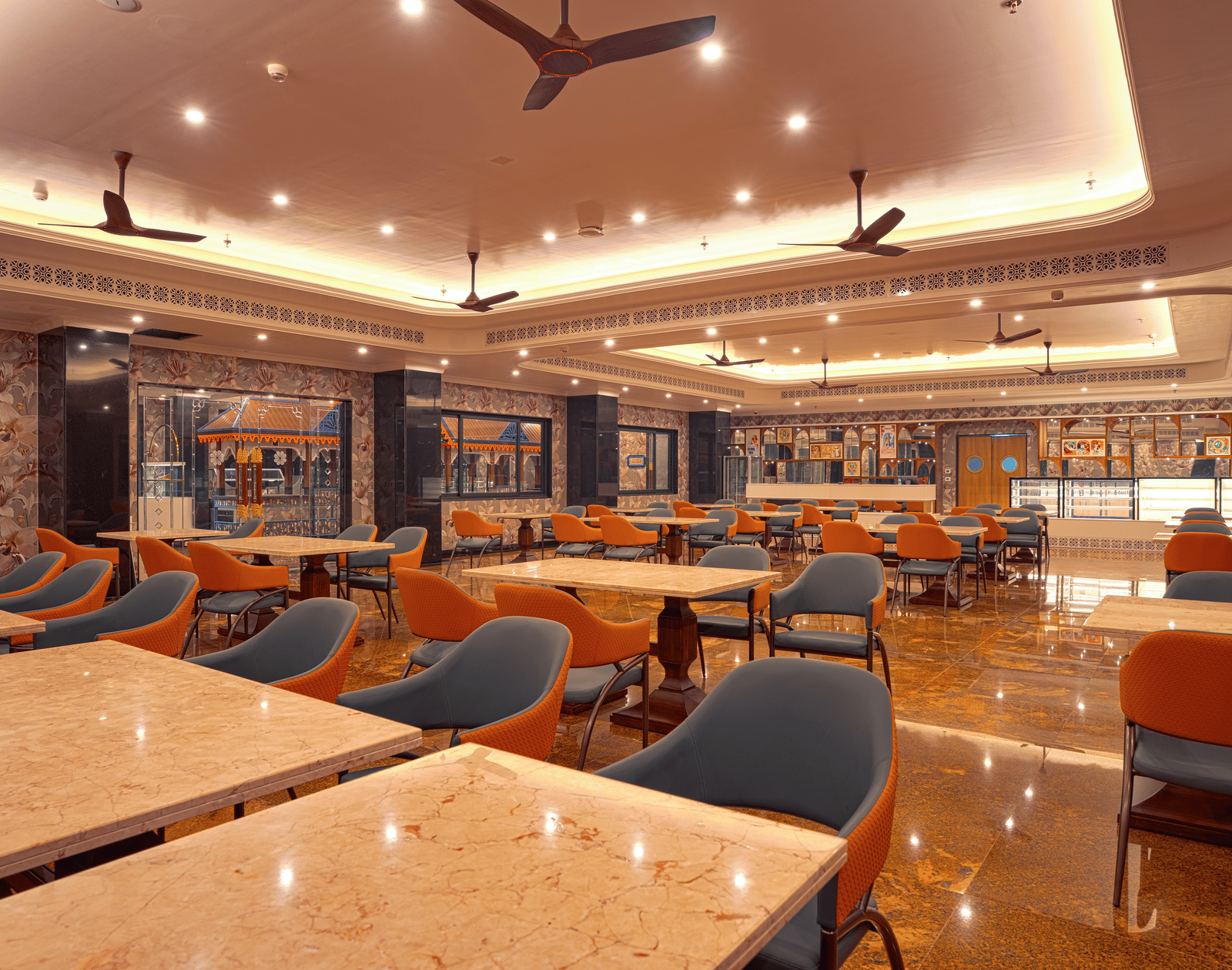 Interior shot of a formal dining room with square marble-topped tables, comfortable teal and orange upholstered chairs, and ceiling fans at MAYFAIR Bay Resort, Paradeep.