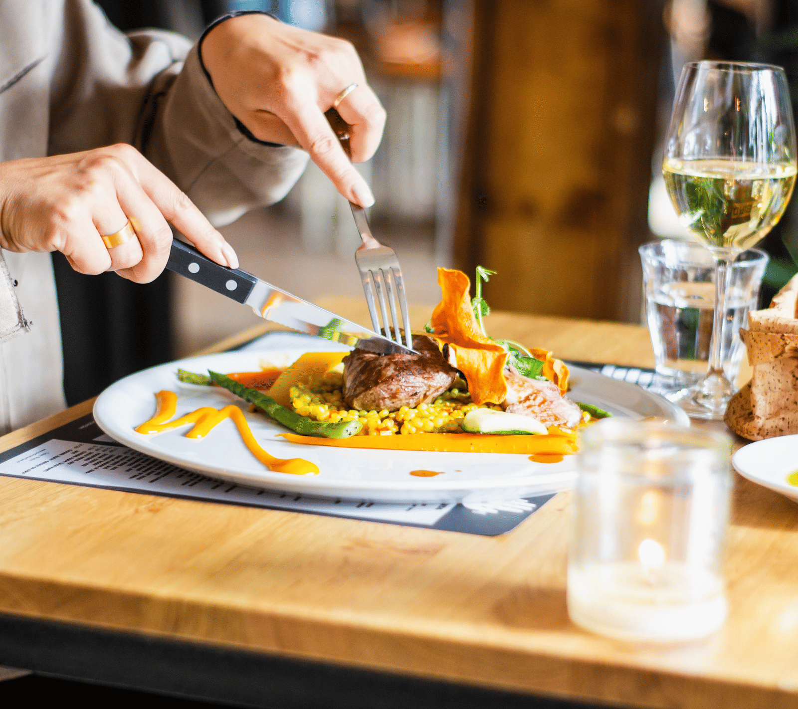 A person cuts into a gourmet meal featuring meat and vegetables on a white plate, accompanied by a glass of white wine and a lit candle on a wooden table.