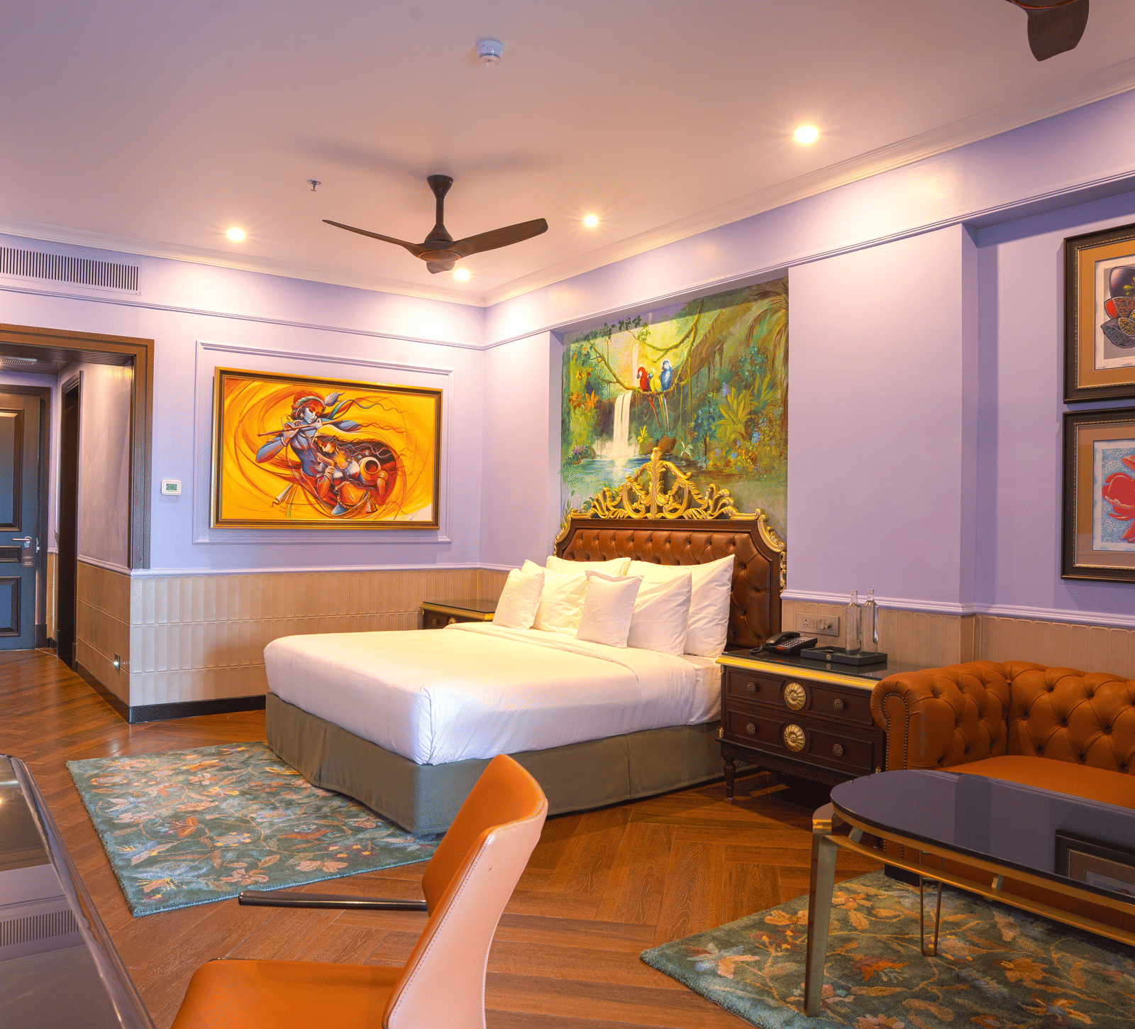 An image of the Executive King room at MAYFAIR Bay Resort, Paradeep. The room features vibrant purple walls, a large bed with white linens, a ceiling fan, colorful artwork including one above the headboard, a patterned teal area rug, and an orange/teal tuf