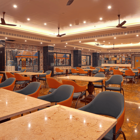 Interior shot of a formal dining room with square marble-topped tables, comfortable teal and orange upholstered chairs, and ceiling fans at MAYFAIR Bay Resort, Paradeep.