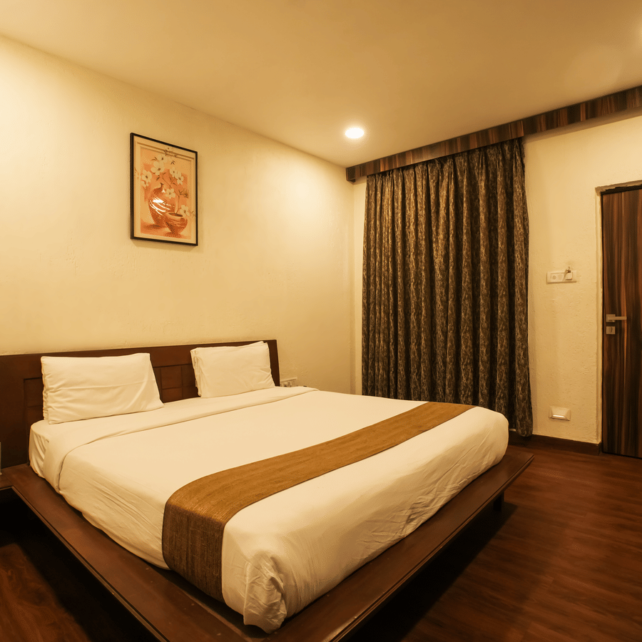 Hotel bedroom featuring a double bed with crisp white sheets, dark wood flooring, and heavy window drapes.