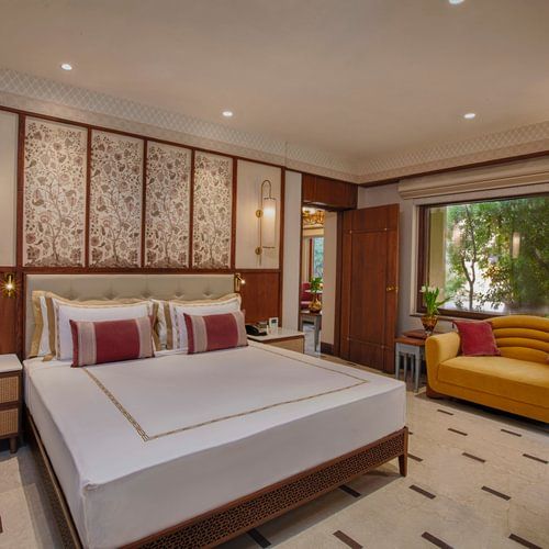 A beautifully designed bedroom with a plush king size bed, elegant floral touches, a cheerful yellow sofa, and a large window offering serene views of lush greenery - Heritage Village Resorts & Spa, Manesar.